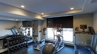 This fitness room is designed to provide a comprehensive workout environment within a compact, efficiently organized space. The room is outfitted with a selection of cardio equipment, including an elliptical trainer, a stair climber, and at least one upright exercise bike, all positioned to take advantage of the natural light streaming through large windows along one wall. The windows are fitted with roller shades, allowing for light control during workouts. On the opposite side of the room, floor-to-ceiling mirrors visually expand the space and allow users to monitor their form during exercise. A well-stocked dumbbell rack lines the mirrored wall, offering a range of free weights alongside medicine balls for varied strength training. The flooring is a durable, dark rubber material suited for high-impact use. Recessed lighting in the ceiling provides consistent, even illumination throughout the room. The overall design is clean and functional, with neutral-toned walls and a practical layout that maximizes usable floor space while maintaining an organized, accessible workout environment.