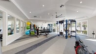 Located at 5724 Owens Dr in Pleasanton, CA, this fitness center offers a comprehensive, single-floor workout facility designed to accommodate a wide range of exercise needs. The space is defined by its bright, open layout, anchored by large floor-to-ceiling windows and glass doors that flood the interior with natural light and provide views of an outdoor pool area and surrounding landscaping.

The flooring features a distinctive striped carpet design in neutral grey tones, lending a clean, contemporary aesthetic throughout. Vaulted and tray ceilings with recessed lighting and large ceiling fans ensure the space remains well-lit and well-ventilated during workouts.

The facility is thoughtfully zoned across its open floor plan. A dedicated cardio section houses multiple treadmills, stair climbers, ellipticals, and stationary bikes from Life Fitness. Adjacent to this area, a strength training zone features a robust blue multi-function cable and functional training rig, along with a full dumbbell rack, weight benches, and a Smith machine. A functional fitness corner is stocked with stability balls, resistance bands, kettlebells, and a rowing machine, catering to dynamic and group training routines.

Wall-mounted mirrors enhance the sense of space and support proper form during exercise. The overall ambiance is clean, modern, and inviting, making this fitness center a well-rounded amenity.
