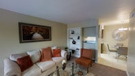 This single-floor space offers a comfortable and well-appointed layout designed for everyday living. The living area serves as the central hub, finished in warm neutral tones with beige walls and carpeted flooring that contribute to an inviting atmosphere. A cream-upholstered sofa dressed with rust-orange accent pillows anchors the seating arrangement, complemented by a cognac-colored accent chair and a glass-top coffee table that add visual depth without overwhelming the space.

A standout feature is the built-in white shelving unit, which provides both display and storage opportunities while acting as a natural divider between the living area and the kitchen beyond. The kitchen is partially visible through this opening, revealing a tiled backsplash, a window that brings in natural light, and functional counter space.

Wall art featuring a scenic wooden bridge adds a touch of nature-inspired décor, reinforcing the warm, earthy color scheme throughout. A glimpse of an additional room to the right suggests a fireplace or decorative feature, adding architectural interest. Overall, the space presents a cohesive, casual aesthetic with practical design elements throughout.