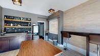 This single-floor communal space blends industrial-modern design with warm, inviting details to create a functional and comfortable gathering area. The space is anchored by a dedicated coffee bar, where rustic wooden open shelving is mounted against a bold chalkboard wall bearing coffee-themed signage. Rich dark cabinetry with granite countertops provides ample counter space, complemented by a professional coffee machine and a water dispenser station nearby. Light hardwood flooring runs throughout, adding warmth against the cool grey walls and crisp white ceilings fitted with stylish cage-style flush-mount light fixtures. A textured stone-tile accent wall adds visual depth to the bar seating area, where a dark bar-height counter and adjustable leather stools offer a casual spot to work or socialize. The lounge area is bathed in natural light from generously sized windows overlooking lush greenery, creating an airy and welcoming atmosphere. A high-top communal table with industrial metal stools sits at the center of the main gathering area, while a plush blue accent chair and a rustic wood coffee table define a cozy lounge nook. A wall-mounted display mount and a secondary room accessible through a doorway round out the functional offerings of this thoughtfully designed communal space.