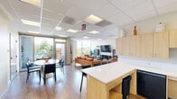 Located at 1490 7th St NW in Washington, DC's Shaw neighborhood, this single-floor community lounge offers a thoughtfully designed, multifunctional space that balances comfort and practicality. The open layout is unified by warm wood-look flooring and crisp white walls, creating a bright and welcoming atmosphere throughout.

The lounge area serves as the focal point of the space, anchored by a set of rich cognac leather sofas and a matching armchair arranged around a sleek oval coffee table. A wall-mounted flat-screen television is flanked by dark display cabinets and an electric fireplace, adding a cozy, living room feel. Lush indoor plants are strategically placed to bring life and warmth to the interior.

Adjacent to the lounge, a casual dining area features a round table with dark chairs positioned near floor-to-ceiling windows that frame views of the surrounding greenery and outdoor spaces, flooding the room with natural light. A bulletin board on the nearby wall serves as a practical community information hub.

The kitchenette is efficiently laid out with light wood cabinetry, a white countertop, a sink, and a refrigerator. A white high-top island with vibrant blue barstools provides additional seating and a casual gathering spot. A set of double doors offers convenient access, completing this versatile and inviting community space.