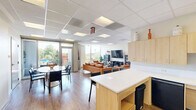 Located at 1490 7th St NW in Washington, DC's Shaw neighborhood, this single-floor community lounge offers a thoughtfully designed, multifunctional space that balances comfort and practicality. The open layout is unified by warm wood-look flooring and crisp white walls, creating a bright and welcoming atmosphere throughout.

The lounge area serves as the focal point of the space, anchored by a set of rich cognac leather sofas and a matching armchair arranged around a sleek oval coffee table. A wall-mounted flat-screen television is flanked by dark display cabinets and an electric fireplace, adding a cozy, living room feel. Lush indoor plants are strategically placed to bring life and warmth to the interior.

Adjacent to the lounge, a casual dining area features a round table with dark chairs positioned near floor-to-ceiling windows that frame views of the surrounding greenery and outdoor spaces, flooding the room with natural light. A bulletin board on the nearby wall serves as a practical community information hub.

The kitchenette is efficiently laid out with light wood cabinetry, a white countertop, a sink, and a refrigerator. A white high-top island with vibrant blue barstools provides additional seating and a casual gathering spot. A set of double doors offers convenient access, completing this versatile and inviting community space.