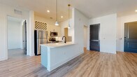 Located at 1440 Lakefront Dr in Sarasota, Florida, this single-story residence showcases a contemporary design with a bright, open-concept layout. Light wood-tone flooring flows throughout the main living areas, complementing the crisp white walls and creating a clean, airy aesthetic.

The expansive living area is generously proportioned, filled with natural light from multiple windows that overlook the surrounding community. A ceiling fan anchors the space, adding both function and a modern accent.

The kitchen is well-appointed with white shaker-style cabinetry featuring matte black hardware, stainless steel appliances including a refrigerator, range, and over-the-range microwave, and a white tile backsplash. A peninsula with a white countertop and black matte faucet provides additional prep space and serves as a casual dining area, illuminated by stylish gold-accented pendant lights. A built-in wine rack adds a distinctive touch to the cabinetry.

The bedroom offers a calm, neutral retreat with natural light from dual windows and the same wood-tone flooring. The bathroom features large-format marble-look tile flooring and clean white walls with matte black hardware accents. A dedicated walk-in storage closet with wire shelving and matching marble-look tile completes the space.