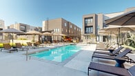 Located at 125 S Navajo St in Salt Lake City, Utah, this single-floor amenity facility spans a generous footprint designed to deliver a comprehensive lifestyle experience. The fitness center is a standout feature, boasting soaring ceilings with exposed industrial ductwork, large floor-to-ceiling windows that flood the space with natural light and frame views of the surrounding landscape, and a wide array of cardio and strength training equipment including treadmills, ellipticals, stationary bikes, rowing machines, and cable machines. Overhead fans and pendant lighting contribute to the functional yet polished aesthetic. Adjacent to the fitness center, a stylishly appointed lounge area features warm terracotta-toned walls, polished concrete floors, high ceilings, curated artwork, and eclectic modern furnishings including plush curved sofas and a sculptural chandelier, creating an inviting social environment. The outdoor amenity area is equally impressive, offering a resort-style lap pool lined with lounge chairs and shaded umbrellas, a covered hot tub with Adirondack seating, and a manicured green lawn bordered by black iron fencing. The covered patio zone provides additional seating for outdoor gatherings, all set against the backdrop of the surrounding modern multi-story residential buildings.