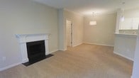 This single-floor space presents a clean, traditional aesthetic characterized by neutral sage-green walls, white crown molding, and white baseboards that frame the interior cohesively. The main living area is anchored by a classic fireplace with a white painted mantel and black surround, serving as a prominent focal point. Plush, light beige wall-to-wall carpeting extends throughout, adding warmth and comfort underfoot. The open layout allows the living area to flow naturally into an adjacent dining or secondary living space, which is illuminated by a simple, modern drum pendant light fixture hanging from the ceiling. Crown molding continues throughout both areas, reinforcing a consistent, polished architectural detail. A white paneled door leads to an additional room or hallway, suggesting further functional space beyond. The overall ambiance is calm and understated, with the neutral color palette and traditional architectural elements creating a versatile backdrop well-suited to a variety of interior design styles. The space feels well-proportioned and move-in ready.