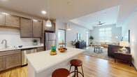 This single-floor residence at 1850 Summerfield Way in Sun Prairie, Wisconsin, offers a thoughtfully designed open-concept layout that seamlessly connects the kitchen, dining, and living areas. The kitchen is a standout feature, equipped with rich, dark-toned cabinetry, a classic subway tile backsplash, stainless steel appliances including a refrigerator, dishwasher, and range, and a large center island topped with a bright white quartz surface. Pendant lighting overhead adds a modern touch, while light hardwood flooring flows throughout the main living spaces. The expansive living room is bathed in natural light from oversized windows and features soaring ceilings with a ceiling fan, creating an airy and welcoming atmosphere with views of the surrounding neighborhood. A secondary carpeted room with wide windows serves as a flexible space, ideal for a den or additional sitting area. The primary bedroom is generously sized with plush carpet, a tray ceiling, and direct access to a well-appointed en-suite bathroom. The bathroom includes a clean, white soaking tub and shower combination with contemporary chrome fixtures. Throughout the residence, the neutral color palette, recessed lighting, and quality finishes contribute to a polished, modern aesthetic.