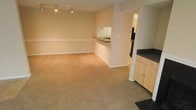 This single-floor space features an open-concept layout that seamlessly connects the living area and kitchen, creating a versatile and functional environment. The living area is finished with wall-to-wall carpeting in a warm neutral tone, contributing to a comfortable and understated ambiance. Walls are painted in a soft beige, maintaining a clean and cohesive aesthetic throughout.

A defining architectural feature is the pass-through counter that separates the kitchen from the living space, providing both visual connectivity and practical utility. The kitchen is equipped with light-toned cabinetry paired with dark countertops, offering a subtle contrast. A sink is visible at the pass-through, and additional counter space and cabinetry extend along the kitchen walls. Dark tile flooring in the kitchen distinguishes it from the carpeted living area.

Track lighting is mounted on the ceiling of the living area, providing adjustable and directed illumination. White chair rail molding runs along the lower portion of the living room walls, adding a traditional architectural detail. The overall layout is open and uncluttered, lending the space a sense of roominess and practicality suitable for everyday living.