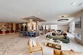 Located at 6276 Newton Rd in Haslett, Michigan, this expansive single-floor community space offers a generously sized open-plan layout designed for both social gathering and relaxed lounging. The interior showcases a thoughtfully curated eclectic modern aesthetic, anchored by striking terrazzo flooring that flows seamlessly throughout the entire space.

The living area features multiple distinct seating zones, including plush sofas and mid-century modern accent chairs arranged around a dramatic arched fireplace nook clad in deep blue tile with built-in shelving. A bold circular pendant light anchors the central lounge area, while floor-to-ceiling windows and sliding glass doors flood the space with natural light and provide direct views of the outdoor patio.

A fully appointed bar and counter area with bar-height seating sits beneath a decorative wood-paneled ceiling inset with globe pendant lights, adding warmth and visual interest. Eclectic wall treatments, including exposed brick, geometric patterned wallcoverings, and curated artwork, create rich textural contrast throughout. Additional zones include dining areas with mixed seating, a billiards table, and intimate conversation nooks. Large windows on multiple walls maintain a bright, welcoming ambiance throughout this well-appointed communal retreat.
