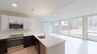 This single-floor unit showcases a clean, contemporary design throughout. The open-concept kitchen and living area serves as the centerpiece, featuring a large island with a white quartz countertop, an undermount sink, and pendant lighting overhead. The kitchen is outfitted with a two-tone cabinetry scheme — dark espresso lower cabinets contrast sharply with crisp white upper cabinets — and includes stainless steel appliances such as a refrigerator, range, microwave, and dishwasher. Light wood-style flooring runs through the kitchen, transitioning into the living area.

The living space is defined by an expansive wall of floor-to-ceiling windows that flood the interior with natural light and provide open views of the surrounding urban skyline, including neighboring brick and high-rise buildings. Recessed lighting throughout reinforces the bright, airy atmosphere.

The bathroom maintains the unit's modern aesthetic with large-format dark tile flooring, beige tile surround in the tub/shower combination, and a dark wood vanity with a white countertop and chrome fixtures. The overall design palette — white walls, neutral tones, and contemporary finishes — creates a cohesive, minimalist ambiance across the entire space.