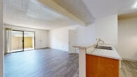 Located in La Mesa, California, this single-story unit offers a practical and functional layout across a well-organized floor plan. The living area features wood-style hard-surface flooring and connects seamlessly to the kitchen, which is partially separated by a peninsula counter with a built-in sink and speckled stone-style countertops. Warm-toned wood cabinetry lines the kitchen walls, complemented by a white electric range with overhead ventilation. A textured popcorn ceiling runs throughout the main living areas, adding a characteristic architectural detail. Natural light flows into the living room through a large sliding glass door that opens to a private patio space enclosed by a wood-panel fence. The bedrooms are carpeted and receive natural light through multiple windows offering outdoor views of surrounding greenery. Two bathrooms are present in the unit; one features a single-sink vanity with matching stone-style countertops and a shower with glass enclosure, while the second offers a wider double-sink vanity and a shower-tub combination. The overall layout is clean, bright, and straightforward, with white walls and neutral finishes throughout providing a versatile backdrop.