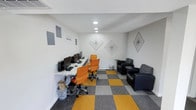 This single-floor space is designed as a functional and modern shared workspace that balances productivity with casual comfort. The room features clean white walls adorned with geometric diamond-shaped line art, lending a contemporary aesthetic to the interior. Recessed ceiling lighting provides even, ambient illumination throughout the space.

The workstation area consists of a long, light-colored desk positioned along one wall, equipped with multiple desktop computers and orange ergonomic office chairs, making it well-suited for individual work or collaborative tasks. A motivational "Start Doing" phrase is displayed on the wall above, reinforcing the workspace's purpose-driven atmosphere.

Adjacent to the workstation area, a lounge section features dark upholstered armchairs arranged around a small side table, offering a comfortable spot for informal meetings or relaxed conversation. The floor is covered in a bold, checkered carpet pattern of alternating mustard yellow and grey tiles, adding a distinctive visual element that ties the two zones together. Overall, the space presents a clean, organized, and creatively styled environment suited for focused work and casual interaction.