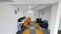 This single-floor space is designed as a functional and modern shared workspace that balances productivity with casual comfort. The room features clean white walls adorned with geometric diamond-shaped line art, lending a contemporary aesthetic to the interior. Recessed ceiling lighting provides even, ambient illumination throughout the space.

The workstation area consists of a long, light-colored desk positioned along one wall, equipped with multiple desktop computers and orange ergonomic office chairs, making it well-suited for individual work or collaborative tasks. A motivational "Start Doing" phrase is displayed on the wall above, reinforcing the workspace's purpose-driven atmosphere.

Adjacent to the workstation area, a lounge section features dark upholstered armchairs arranged around a small side table, offering a comfortable spot for informal meetings or relaxed conversation. The floor is covered in a bold, checkered carpet pattern of alternating mustard yellow and grey tiles, adding a distinctive visual element that ties the two zones together. Overall, the space presents a clean, organized, and creatively styled environment suited for focused work and casual interaction.