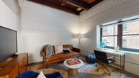 This single-floor space showcases a distinctive loft-inspired aesthetic, defined by its signature white-painted exposed brick walls and rich, dark wood beam ceilings that lend an industrial yet refined character throughout. Exposed ductwork adds an authentic urban edge, while the open layout creates a naturally flowing living environment.

The living area is generously proportioned, anchored by a cognac leather sofa, a plush navy accent chair, and a marble-topped coffee table set atop a layered vintage-style rug. A wood media console houses a flat-screen television, completing the comfortable and stylish entertaining setup. A floor lamp and a window providing natural light enhance the warm ambiance of the room.

The kitchen features white upper cabinetry contrasted by dark lower cabinets, a dark tile backsplash, and stainless steel appliances including a range, dishwasher, and a full-size refrigerator. Track lighting illuminates the space efficiently. An adjacent dining table with seating integrates seamlessly into the kitchen area.

The bedroom offers a calm, minimalist retreat with smooth white walls, exposed beam ceiling details, a pendant light, and a cozy area rug. The bathroom is clean and contemporary, featuring a dark vanity with an integrated sink, tile flooring, and a shower with a curtain enclosure.