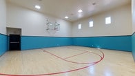 This dedicated indoor basketball court is a well-appointed athletic space designed for serious recreational play. The court features a beautifully finished light maple hardwood floor with crisp red boundary lines, lane markings, and a painted arc, all consistent with standard court layout. A wall-mounted glass backboard and hoop are positioned at one end, providing a professional-grade playing experience. The lower portion of the walls is painted in a bold teal accent band, adding a dynamic visual contrast against the clean white upper walls and ceiling. Recessed skylight-style flush ceiling fixtures flood the court with bright, even illumination, ensuring excellent visibility throughout the space. A row of upper-level windows along the side wall allows natural light to filter in, enhancing the overall atmosphere. The high ceiling provides ample vertical clearance for unobstructed gameplay. The space is clean, functional, and purpose-built, making it an ideal facility for casual pick-up games, skill training, or organized play. The combination of quality flooring, proper court markings, and thoughtful lighting design reflects a commitment to delivering a premium indoor sports environment.