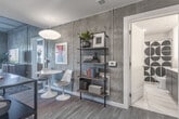 Located at 111 Hull St in Richmond, Virginia, this contemporary single-floor residence blends modern design with urban sophistication. The kitchen is outfitted with dark espresso cabinetry, stainless steel appliances including a gas range and over-the-range microwave, and a clean white countertop that contrasts elegantly against the bold cabinetry. A sliding glass door at the end of the open-concept living and dining area floods the space with natural light and offers views of the Richmond skyline, creating a bright and inviting atmosphere. The living area features wood-look flooring, a tufted light gray sofa, a geometric-patterned area rug, and contemporary pendant lighting, all contributing to a cohesive, stylish aesthetic. The bedroom is a standout retreat, distinguished by a dramatic textured dark accent wall, large windows framing sweeping city and treetop views, hardwood-style flooring, and curated art pieces. A warm wood dresser and a vibrant orange accent chair add character to the space. The bathroom is finished with large-format marble-look tile flooring, a dark wood vanity with backlit mirror, and direct access to a spacious walk-in closet with wire shelving. Throughout the residence, thoughtful design details and quality finishes create a refined, urban living experience.