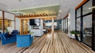 Located at 131 E Green St in Ithaca, NY, this generously sized single-floor amenity space offers a thoughtfully designed environment across several distinct areas. The main lounge serves as the heart of the space, featuring warm wood-beam pergola accents, a large wall-mounted TV with a built-in fireplace below, and an assortment of stylish seating including plush sofas and bold blue velvet accent chairs. Floor-to-ceiling glass walls and expansive windows flood the area with natural light while providing views of the surrounding streetscape. Sleek bar-height tables complement the open layout, creating versatile gathering spots. Adjacent to the lounge, a dedicated game and recreation room features a full-size pool table, a beverage station, and eye-catching geometric-patterned accent walls in deep teal and silver. The fitness center is well-equipped with Peloton cycling bikes, a multi-station cable machine, a full dumbbell rack, and mirrored walls, all set on durable rubberized flooring beneath bright overhead lighting. Black-framed glass partition walls define separate office or collaborative spaces, offering transparency while maintaining privacy. A connecting hallway with wood-look flooring and colorful accent walls ties the areas together, providing access to additional rooms and facilities throughout the floor.