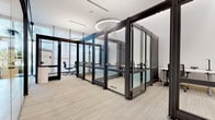 This contemporary single-floor office space is thoughtfully designed to balance open collaboration with private, focused work. The layout is defined by a series of modular glass-and-metal partition systems featuring dark-framed enclosures with full-height glazing, creating individual workstations and enclosed meeting rooms while maintaining strong visual connectivity throughout the floor.

The workstation pods are equipped with adjustable-height desks and ergonomic task chairs, offering a functional and flexible environment for individual productivity. Adjacent enclosed rooms accommodate small group meetings, furnished with round conference tables and sleek leather chairs. A whiteboard wall panel is visible within one meeting room, supporting collaborative sessions.

Flooring throughout consists of light-toned, textured carpet tiles that complement the neutral white ceiling and walls. Overhead, striking circular pendant light fixtures provide ambient illumination, adding a refined architectural element to the space. Natural light filters in through expansive exterior glazing, with views of palm trees suggesting a warm, sunny locale.

Warm wood-paneled accent walls feature prominently in certain enclosures, introducing a natural contrast to the otherwise cool, polished material palette. The overall design conveys a professional, well-appointed workspace suited for modern business operations.