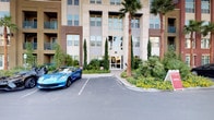 This impressive building presents a polished and welcoming first impression through its beautifully landscaped exterior, featuring tall palm trees, Italian cypress, and vibrant flowering shrubs framing a paved entry courtyard. The facade is finished in warm stone and neutral tones, with large windows spanning multiple levels that allow natural light to permeate throughout. A set of sleek glass double doors opens into a dramatic lobby space defined by soaring double-height ceilings and an eye-catching sputnik chandelier that anchors the room with a bold contemporary flair. The lobby is finished with wide-plank wood-look flooring that runs seamlessly throughout, lending warmth and continuity to the space. A large-format mural depicting a stunning canyon landscape commands one wall, creating a vivid focal point that adds artistic character to the interior. The reception desk, finished in dark cabinetry with an illuminated stone countertop, serves as a refined and functional centerpiece. Recessed cove lighting along the ceiling adds a soft, layered glow, complementing the pendant lights above the reception area. A mezzanine-level overlook with black metal railings adds architectural depth, while a lounge area visible in the background invites further exploration of the space.