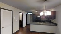 This single-floor space showcases a contemporary open-concept layout that seamlessly connects the kitchen and living areas. The kitchen is a standout feature, equipped with dark cabinetry, a black refrigerator, and a peninsula countertop with a granite-style surface and an integrated sink — ideal for both meal preparation and casual dining. Overhead, a decorative multi-bulb pendant light fixture adds a modern industrial touch, while a track lighting system provides additional illumination across the space. Rich, dark hardwood flooring runs throughout, creating a warm yet sophisticated contrast against the light-toned walls. A transom window above the entryway allows natural light to filter in near the front door, which features a contemporary panel design and a keypad entry system for added security. A hallway off the main living area leads to a bathroom, which is partially visible and includes a vanity with a mirror and dark cabinetry consistent with the overall design aesthetic. A programmable thermostat is mounted on the wall, reflecting the unit's thoughtful, functional design. The overall ambiance is clean, modern, and well-appointed, with cohesive dark accents that lend a polished, urban character to the space.