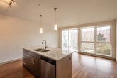 This single-floor space showcases a thoughtfully designed open-concept layout that seamlessly connects the kitchen and living area, creating a bright and functional living environment. Warm hardwood flooring flows consistently throughout, lending a sense of continuity and warmth to the entire space.

The kitchen is a standout feature, outfitted with rich dark wood cabinetry, stainless steel appliances including a refrigerator, range, and built-in microwave, and a generous island topped with elegant granite countertops. A stainless steel undermount sink is integrated into the island, which also houses a dishwasher. Pendant lighting and a track light system illuminate the kitchen, while a mosaic tile backsplash adds a refined decorative touch. A large window at the far end of the living area floods the space with natural light and provides views of surrounding greenery.

A narrow hallway connects the main living area to the private spaces, featuring a sliding door and a ceiling-mounted light fixture. The bathroom is cleanly finished with a dark wood vanity topped with a marble-style countertop, hardwood-style flooring, and a full bathtub and shower combination with a curved curtain rod, all set against crisp white walls and fixtures. The overall ambiance is clean, contemporary, and move-in ready.