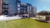 This contemporary multi-story residential community showcases a striking exterior defined by a modern architectural design with clean lines, contrasting grey and blue paneling, and red accent elements. The building rises several stories, with each level featuring private balconies that offer residents outdoor access and views of the surrounding area.

At the ground level, the community centers around a thoughtfully designed outdoor courtyard that serves as a social and recreational hub. A generous stretch of lush green turf provides an open, inviting lawn area ideal for relaxation and outdoor activities. Adjacent to the lawn, a spacious patio area is furnished with comfortable navy blue seating arrangements, creating an outdoor living room atmosphere perfect for gathering and lounging.

The courtyard is further enhanced by a covered pergola structure, additional seating clusters, and umbrellas that provide shade throughout the day. Tasteful landscaping, including palm trees and other plantings, adds a warm, resort-like character to the space. Tall exterior light poles ensure the outdoor areas remain well-lit in the evenings.

The entire outdoor space benefits from abundant natural sunlight and open skies, contributing to a bright and welcoming ambiance. The cohesive design and well-maintained grounds reflect a strong attention to detail, making this community's outdoor amenity space both functional and visually appealing.