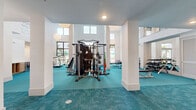 Located at 9005 Solay Rd in Winter Garden, Florida, this well-appointed fitness center offers an expansive, open-concept layout across a single floor. The space is defined by its dramatic double-height ceilings, which flood the interior with natural light through large floor-to-ceiling windows and elevated clerestory windows along the upper walls. Crisp white walls and structural columns are complemented by striking teal carpet tile flooring, giving the space a vibrant and energizing aesthetic. Decorative woven pendant lights add a refined touch overhead, while motivational quotes are displayed along the upper walls.

The fitness floor is comprehensively equipped for a full-body workout experience. A central multi-station cable and weight machine anchors the strength training area, surrounded by free weight racks, adjustable benches, a punching bag, and a plyometric box. The cardio zone features treadmills, a stair climber, and an air resistance bike, all positioned near windows overlooking lush Florida greenery. A built-in beverage cooler provides convenient refreshment access. Wall-mounted mirrors enhance the sense of space and support training feedback. A sanitation station with towels and supplies is also present, ensuring a clean and well-maintained environment throughout.