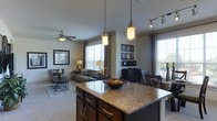 This single-story space showcases an open-concept layout that integrates the kitchen, dining, and living areas into one cohesive environment. The kitchen features a large center island topped with a speckled granite countertop and dark cabinetry with brushed gold hardware. Pendant lights hang above the island, providing warm, focused illumination that complements the track lighting mounted on the ceiling. The living area is carpeted and receives generous natural light through multiple large windows dressed with layered curtains and blinds. A ceiling fan with a light fixture serves the living space, adding both function and a finishing touch. The dining area sits adjacent to the living room, furnished with dark wood chairs and a matching table. A display cabinet with decorative framed artwork adds visual interest to the space. The overall color palette is neutral, with warm tones created by the cabinetry, granite, and lighting fixtures. The open flow between spaces makes the layout feel expansive and practical, offering a comfortable and well-appointed environment suitable for everyday living and entertaining.