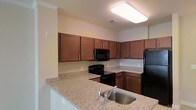 This single-floor residence offers a thoughtfully designed living environment with a kitchen that serves as a central highlight of the space. The kitchen is outfitted with warm-toned wood cabinetry that provides generous storage, complemented by light granite-style countertops that extend across the workspace and into a convenient peninsula or breakfast bar area. A stainless steel sink is positioned along the counter for easy accessibility. The suite of black appliances — including a refrigerator, electric range, and over-the-range microwave — creates a unified, classic aesthetic throughout the kitchen. Crown molding accents the upper walls, adding a refined architectural touch. A large flush-mounted ceiling light fixture ensures the kitchen is well-lit and functional. The neutral, warm wall tones throughout contribute to an inviting and comfortable ambiance. The layout of the kitchen is efficient and practical, with ample counter space for meal preparation and cabinetry arranged to maximize storage. Overall, the space reflects a clean, functional design with quality finishes that offer both style and everyday practicality.