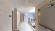 This shared hallway corridor is designed to provide efficient access to multiple units within the building. The space features a textured stucco finish on the walls and ceiling, lending a durable, low-maintenance aesthetic throughout. Smooth concrete flooring runs the length of the corridor, offering a clean and practical surface well-suited to high-traffic use.

A standout architectural element is the open metal railing system along one side of the hallway, which adds a contemporary industrial touch while providing safety along the open edge. The railing overlooks a lower level, reinforcing the multi-story nature of the building. Circular wall-mounted sconce lighting illuminates the corridor evenly, complementing the natural light that filters in through porthole-style windows set into the exterior wall.

Unit entry doors are positioned along the hallway, each clearly numbered for easy navigation. An illuminated exit sign above one of the doors ensures clear emergency wayfinding. The light blue accent wall at the corridor's end adds a subtle visual contrast against the surrounding neutral tones, contributing to an overall atmosphere that is clean, functional, and straightforward.