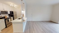 This single-story, 2-bedroom, 1-bathroom residence is designed with a clean, contemporary aesthetic defined by a neutral white and light wood palette throughout. The open-concept living and dining area is anchored by light wood-style flooring and flows seamlessly toward a sliding glass door dressed with vertical blinds, allowing abundant natural light to pour in while providing access to an outdoor view. A built-in shelving unit adds a functional decorative element to the living space.

The kitchen is positioned as a peninsula-style layout, separated from the main living area by a white countertop bar that doubles as a casual dining surface. White cabinetry, stainless steel appliances including a refrigerator and microwave, and pendant lighting over the peninsula contribute to the kitchen's modern and practical design. Track lighting illuminates the galley-style cooking corridor.

A central hallway connects the two bedrooms, both of which feature plush carpeting, ceiling fans, and their own sliding glass doors that invite natural light and outdoor views. The shared full bathroom is well-appointed with white cabinetry, a wide vanity with ample storage, a large mirror, and a tub-shower combination.