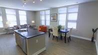This single-floor space is designed with a seamless open-concept layout that connects the kitchen, dining, and living areas into one cohesive, functional environment. The kitchen features a dark countertop island that doubles as a visual divider, complemented by stainless steel appliances and easy access to both the dining and living spaces. The dining area accommodates a dark wood table paired with cream upholstered chairs, adding a refined contrast that anchors the center of the space. The living area beyond provides a comfortable lounge setting, accented by a bold orange armchair that adds a vibrant pop of color against the neutral gray walls. Hardwood-style flooring flows consistently throughout, lending warmth and visual continuity across the entire space. Multiple large windows dressed with horizontal blinds line the walls, inviting generous amounts of natural light and creating an airy, open ambiance. Recessed track lighting along the ceiling provides additional illumination, ensuring the space is well-lit at all times. A framed piece of wall art adds a personal decorative touch. The overall aesthetic is modern and clean, blending neutral tones with warm accents for a balanced and welcoming atmosphere.