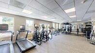 Located at 12 Valley St in Everett, MA, this fitness center offers a well-equipped, single-floor exercise facility designed to support a full range of workout routines. The open layout is finished with neutral-toned carpet throughout the cardio zone and rubber flooring in the strength training area, providing both comfort and functionality underfoot.

The cardio section is lined along the windowed wall, where multiple treadmills, elliptical trainers, and a stationary bike are arranged to take advantage of the natural light streaming in through several large windows. The brightness of the space is further enhanced by recessed panel lighting set into a drop-tile ceiling, keeping the environment well-lit and inviting.

The strength training area is equally comprehensive, featuring a multi-station cable machine, a power rack with bench press capability, and a well-stocked dumbbell rack with a full range of free weights. Full-length mirrors line one wall of this section, amplifying the sense of space and allowing users to monitor their form during workouts.

The facility also includes a designated hand sanitizer station and informational signage, reflecting a commitment to cleanliness and user awareness. With its thoughtful equipment arrangement, natural light, and clean, modern aesthetic, this fitness center provides a practical and motivating environment for residents seeking a convenient on-site workout space.