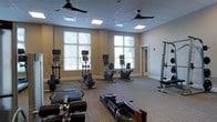 This single-floor fitness center is designed to provide a comprehensive workout environment in a clean, well-organized space. The room features large windows along one wall, flooding the interior with natural light and offering views of surrounding greenery, creating an open and inviting atmosphere. A drop-panel ceiling with recessed lighting and multiple ceiling fans ensures consistent illumination and airflow throughout the space.

The flooring consists of durable, patterned carpet tiles suited for a high-traffic fitness environment. Cardio equipment is prominently featured, including several upright and recumbent stationary bikes and elliptical machines, each equipped with individual monitors for entertainment or workout tracking. Rowing machines are positioned toward the front of the room, offering a full-body workout option. A vertical weight storage rack along the side wall provides organized access to free weights.

The neutral, warm-toned walls complement the modern equipment and contribute to a focused yet comfortable workout setting. A secondary doorway provides additional access or connects to an adjoining room. Overall, the space balances functionality with a clean aesthetic, making it suitable for a wide range of fitness routines.