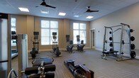This single-floor fitness center is designed to provide a comprehensive workout environment in a clean, well-organized space. The room features large windows along one wall, flooding the interior with natural light and offering views of surrounding greenery, creating an open and inviting atmosphere. A drop-panel ceiling with recessed lighting and multiple ceiling fans ensures consistent illumination and airflow throughout the space.

The flooring consists of durable, patterned carpet tiles suited for a high-traffic fitness environment. Cardio equipment is prominently featured, including several upright and recumbent stationary bikes and elliptical machines, each equipped with individual monitors for entertainment or workout tracking. Rowing machines are positioned toward the front of the room, offering a full-body workout option. A vertical weight storage rack along the side wall provides organized access to free weights.

The neutral, warm-toned walls complement the modern equipment and contribute to a focused yet comfortable workout setting. A secondary doorway provides additional access or connects to an adjoining room. Overall, the space balances functionality with a clean aesthetic, making it suitable for a wide range of fitness routines.