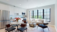 This thoughtfully designed single-floor residence showcases a seamless open-concept layout that integrates the kitchen, dining, and living areas into one cohesive, light-filled space. The living room is anchored by a striking bank of large, black-framed grid windows that flood the interior with natural light and offer views of the surrounding urban streetscape, creating an airy and inviting atmosphere. Warm hardwood flooring runs throughout the main living areas, complementing the neutral grey and white color palette.

The kitchen is outfitted with sleek white cabinetry, stone countertops, a deep undermount sink with a modern gooseneck faucet, a tile backsplash, and stainless steel appliances including a gas range and built-in microwave. A round dining table nestled between the kitchen and living area provides an ideal space for casual meals.

The bedroom is separated from the main living area by an elegant black-framed frosted glass sliding partition, offering privacy while maintaining an open, loft-like feel. The bedroom includes direct access to a full bathroom finished with tile flooring and contemporary fixtures. An additional bathroom serves the rest of the residence, featuring clean, minimal finishes consistent with the overall modern aesthetic of the space.