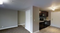 This single-floor residence offers a practical and well-organized layout designed for comfortable everyday living. The living area features neutral-toned carpet flooring and smooth, white-trimmed walls painted in a warm greige, creating a calm and understated atmosphere throughout the space. A closet with white bi-fold doors provides convenient storage directly off the main living area, while a thermostat panel is neatly mounted on the adjacent wall for easy climate control access. The transition from the carpeted living space to the kitchen is marked by a shift to hard surface flooring, lending a clean visual distinction between the two zones. The kitchen is equipped with dark espresso-stained cabinetry that extends to the upper walls, offering ample storage. Black appliances, including an electric range and an over-the-range microwave, complement the dark cabinetry for a cohesive aesthetic. A sink is positioned along the counter, making the kitchen both functional and efficient. The overall design of the space is straightforward and move-in ready, with a neutral color palette that provides a versatile backdrop for a variety of interior styles.