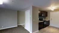 This single-floor residence offers a practical and well-organized layout designed for comfortable everyday living. The living area features neutral-toned carpet flooring and smooth, white-trimmed walls painted in a warm greige, creating a calm and understated atmosphere throughout the space. A closet with white bi-fold doors provides convenient storage directly off the main living area, while a thermostat panel is neatly mounted on the adjacent wall for easy climate control access. The transition from the carpeted living space to the kitchen is marked by a shift to hard surface flooring, lending a clean visual distinction between the two zones. The kitchen is equipped with dark espresso-stained cabinetry that extends to the upper walls, offering ample storage. Black appliances, including an electric range and an over-the-range microwave, complement the dark cabinetry for a cohesive aesthetic. A sink is positioned along the counter, making the kitchen both functional and efficient. The overall design of the space is straightforward and move-in ready, with a neutral color palette that provides a versatile backdrop for a variety of interior styles.
