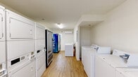 Located at 644 W Magnolia Ave in Auburn, Alabama, this community laundry facility offers a clean, efficiently organized space designed for convenient shared use. The room features light-toned walls and warm hardwood-style flooring that create a bright, welcoming atmosphere throughout. Overhead fluorescent lighting ensures the space is well-lit at all times.

The facility is equipped with multiple top-loading Whirlpool washers and stacked washer-dryer units, providing ample laundry capacity for residents. A dedicated trash receptacle is conveniently positioned near the machines. A Dasani glass-door beverage vending machine adds a practical amenity to the space. A wall-mounted mini-split air conditioning unit keeps the room comfortable year-round.

A window with blinds near the entrance allows for natural light and ventilation, while a keyed entry door with an electronic lock ensures secure access. An electrical panel is recessed into one wall, keeping utility infrastructure neatly integrated. The overall layout is thoughtfully arranged to allow easy movement between machines, making this a functional and user-friendly shared laundry facility.
