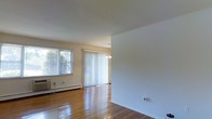 This single-floor space offers a clean and functional layout with a welcoming, understated aesthetic. The living area is defined by gleaming hardwood floors that run throughout, reflecting natural light and adding warmth to the interior. A large window and full-length sliding glass door fitted with vertical blinds flood the space with abundant daylight, creating an airy and open atmosphere. The sliding door provides a direct connection to the outdoors, enhancing the sense of openness. A modest chandelier overhead adds a subtle decorative touch to the dining or transitional area, complementing the otherwise simple and neutral interior. The walls are finished in a clean white tone, offering a versatile backdrop suitable for a variety of uses and furnishing styles. A baseboard heating unit runs along the exterior wall beneath the window, providing efficient climate control. The open flow between areas makes the space feel generously proportioned, with clear sightlines from one end to the other. Overall, the space presents a bright, well-maintained environment with classic hardwood detailing and practical features throughout.