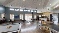 This single-story community clubhouse is thoughtfully designed to accommodate a variety of social and recreational activities. The space is characterized by soaring vaulted and drop ceilings throughout, with clerestory windows lining the upper walls to flood the interior with abundant natural light. Durable wood-look flooring anchors the main areas, complemented by slate-toned walls that lend a modern, cohesive aesthetic.

The expansive game room serves as a central hub, featuring ping-pong and billiard tables alongside a large retractable garage door that opens to the outdoors, creating a seamless indoor-outdoor connection. Multiple seating arrangements are distributed throughout, including a lounge area with leather sofas, accent chairs, and wall-mounted televisions. A bold charcoal wood-paneled accent wall with a decorative logo adds a distinctive focal point to one of the lounges.

The kitchen is well-appointed with granite countertops, a center island, mosaic tile backsplash, white cabinetry, stainless steel appliances, and pendant lighting. A coffee station and dining tables with chairs offer additional amenities for residents. A separate corridor leads to additional rooms, completing this versatile, amenity-rich facility.