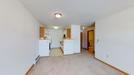 This single-floor space features an open-concept design that integrates the kitchen and living area into one cohesive layout. The kitchen is equipped with white appliances, including a refrigerator and range, complemented by oak-toned cabinetry and light countertops. A peninsula-style divider separates the kitchen from the carpeted living area, offering a natural transition between the two spaces. The kitchen flooring is finished in light-toned tile, providing a practical and easy-to-maintain surface, while the living area features wall-to-wall carpet for a comfortable feel underfoot. Flush-mount ceiling fixtures provide overhead lighting throughout, and the textured ceiling adds a classic finishing detail. A baseboard heater runs along the living area wall, offering an efficient heating solution. White walls throughout create a clean, bright atmosphere. A hallway extending from the main area leads to a bathroom, which is visible at the end of the corridor. The overall layout is functional and straightforward, with a neutral color palette and practical finishes that create a welcoming, low-maintenance environment.