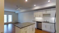 This single-floor space showcases a clean, modern design centered around an open-concept layout that seamlessly blends the kitchen and living areas. The kitchen is a standout feature, outfitted with crisp white shaker-style cabinetry equipped with sleek brushed-nickel hardware, offering generous storage both above and below the counters. Dark granite countertops provide a striking contrast against the white cabinetry, while a matching granite-topped center island serves as a functional focal point, ideal for meal preparation or casual dining. Stainless steel appliances, including a built-in dishwasher, add a contemporary touch. A classic white subway tile backsplash completes the kitchen's polished aesthetic. The flooring throughout is rich hardwood, lending warmth and continuity to the open space. Recessed lighting and a track lighting fixture ensure the space is well-illuminated, creating a bright and inviting atmosphere. The open flow between the kitchen and the adjacent living area enhances the sense of spaciousness, making the layout well-suited for both everyday living and entertaining. The neutral color palette of soft whites and grays contributes to an overall ambiance that feels fresh, airy, and move-in ready.