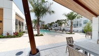 The Residences at Enso presents a thoughtfully designed outdoor amenity space centered around a generous resort-style swimming pool. The pool area is framed by tall, mature palm trees that lend a tropical ambiance, while the surrounding travertine paver deck provides an elegant, durable surface ideal for lounging and entertaining. Rows of chaise lounge chairs and market umbrellas are arranged along the pool's perimeter, offering ample space for residents to relax in the open air.

The pool clubhouse building features a striking contemporary design, combining crisp white panel cladding with rich warm wood siding accents. Large floor-to-ceiling glass doors and windows provide visual connectivity between the interior amenity spaces and the outdoor pool deck. A covered outdoor area with exposed wood-beam ceilings and sturdy timber support columns serves as a shaded retreat, complete with seating that overlooks the water.

Lush, manicured landscaping with tropical plants and ornamental grasses borders the pool, softening the architectural lines and enhancing the resort atmosphere. The broader community buildings visible in the background echo the same modern white and warm-toned aesthetic, creating a cohesive and polished environment. The overall ambiance is that of a refined, upscale outdoor living space designed for both relaxation and community gathering.