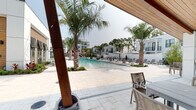 The Residences at Enso presents a thoughtfully designed outdoor amenity space centered around a generous resort-style swimming pool. The pool area is framed by tall, mature palm trees that lend a tropical ambiance, while the surrounding travertine paver deck provides an elegant, durable surface ideal for lounging and entertaining. Rows of chaise lounge chairs and market umbrellas are arranged along the pool's perimeter, offering ample space for residents to relax in the open air.

The pool clubhouse building features a striking contemporary design, combining crisp white panel cladding with rich warm wood siding accents. Large floor-to-ceiling glass doors and windows provide visual connectivity between the interior amenity spaces and the outdoor pool deck. A covered outdoor area with exposed wood-beam ceilings and sturdy timber support columns serves as a shaded retreat, complete with seating that overlooks the water.

Lush, manicured landscaping with tropical plants and ornamental grasses borders the pool, softening the architectural lines and enhancing the resort atmosphere. The broader community buildings visible in the background echo the same modern white and warm-toned aesthetic, creating a cohesive and polished environment. The overall ambiance is that of a refined, upscale outdoor living space designed for both relaxation and community gathering.