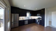 This single-floor space makes a striking impression with its thoughtfully designed kitchen at the heart of the layout. The kitchen showcases floor-to-ceiling dark espresso cabinetry that creates a bold, contemporary aesthetic, contrasted beautifully by crisp white countertops that reflect light and add a sense of brightness to the space. The cabinetry is configured in an L-shaped layout, maximizing storage and providing an efficient workflow for cooking and entertaining. A full suite of stainless steel appliances — including a French door refrigerator, gas range, built-in microwave, and dishwasher — lends a professional, polished finish to the kitchen. Recessed lighting in the white ceiling ensures the space is well-lit, while a window above the sink invites a touch of natural light, enhancing the overall ambiance. Rich, dark hardwood flooring runs throughout, tying together the dramatic color palette and adding warmth to the modern design. The clean lines, minimal hardware, and high-contrast tones throughout give this space a refined, contemporary character that is both functional and visually compelling.