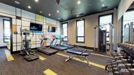 This fitness center is thoughtfully designed to support a full range of workout disciplines across multiple interconnected spaces. The cardio area serves as a focal point, housing a row of treadmills and an elliptical trainer positioned in front of a striking accent wall featuring a colorful geometric grid pattern. A wall-mounted flat-screen television accompanies the cardio equipment, while recessed lighting and a ceiling fan illuminate the space from above a dark-painted ceiling. Natural light enters through multiple dark-framed windows, adding brightness to the interior.

A dedicated cycling studio offers yellow-accented stationary bikes arranged on a raised platform section, complemented by floor-to-ceiling mirrors and an additional mounted television. The room benefits from ample window views to the outdoors. A separate strength training zone provides a comprehensive dumbbell rack, a cable machine system, a kettlebell, and a weight bench, all reflected in large wall mirrors that enhance the sense of space.

Connecting corridors feature bold yellow typographic wall graphics, dark-trimmed doors, and the same striped carpet pattern that unifies the facility. A boxing area with a heavy punching bag and speed bag rounds out the amenity offerings. Throughout, the charcoal, yellow, and white color palette creates an energetic and cohesive aesthetic.