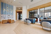 Located at 5851 Columbia Pike in Falls Church, Virginia, this expansive residential lobby spans nearly 2,900 square feet on a single floor, offering a refined and thoughtfully designed first impression. The entrance is anchored by dramatic floor-to-ceiling windows that flood the space with natural light and frame views of the surrounding streetscape, creating an airy, open ambiance from the moment you step inside.

The main lobby area features light-toned hardwood flooring that flows seamlessly throughout, complemented by soaring ceilings and a striking geometric white sculptural wall panel that serves as a bold architectural focal point. A coordinated blue-and-gold reception desk with accompanying upholstered seating in gold wire-frame chairs defines the leasing and welcome area, exuding a contemporary yet warm aesthetic. Adjacent to the reception zone, a glass-enclosed lounge offers a visually connected yet distinct space, complete with pendant lighting and stylish furnishings.

A dedicated mailroom corridor provides residents with convenient access to individual mailboxes, accented by modern pendant light fixtures and clean tile flooring. Throughout the lobby, details such as fluted wall treatments, warm wood accents, and curated pendant chandeliers contribute to an elevated, cohesive design language that balances sophistication with everyday functionality.