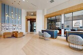 Located at 5851 Columbia Pike in Falls Church, Virginia, this expansive residential lobby spans nearly 2,900 square feet on a single floor, offering a refined and thoughtfully designed first impression. The entrance is anchored by dramatic floor-to-ceiling windows that flood the space with natural light and frame views of the surrounding streetscape, creating an airy, open ambiance from the moment you step inside.

The main lobby area features light-toned hardwood flooring that flows seamlessly throughout, complemented by soaring ceilings and a striking geometric white sculptural wall panel that serves as a bold architectural focal point. A coordinated blue-and-gold reception desk with accompanying upholstered seating in gold wire-frame chairs defines the leasing and welcome area, exuding a contemporary yet warm aesthetic. Adjacent to the reception zone, a glass-enclosed lounge offers a visually connected yet distinct space, complete with pendant lighting and stylish furnishings.

A dedicated mailroom corridor provides residents with convenient access to individual mailboxes, accented by modern pendant light fixtures and clean tile flooring. Throughout the lobby, details such as fluted wall treatments, warm wood accents, and curated pendant chandeliers contribute to an elevated, cohesive design language that balances sophistication with everyday functionality.
