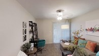 This single-floor space offers a warm and functional layout designed to accommodate both relaxation and productivity. The main living area is finished with soft wall-to-wall carpeting underfoot, contributing to a comfortable and inviting atmosphere. A ceiling fan with a light fixture serves as the central overhead lighting, while an arc floor lamp adds a stylish supplementary glow to the room. A sliding glass door fitted with vertical blinds allows for adjustable natural light and provides a connection to the outdoors, brightening the space throughout the day. The neutral, light-toned walls create a clean and versatile backdrop that complements a range of décor styles. A dedicated workspace area is thoughtfully incorporated into the layout, featuring a leaning bookshelf and a compact desk paired with a teal accent chair, making it well-suited for working from home or studying. Wall-mounted decorative mirrors add visual interest and depth to the room. Lush potted plants and decorative florals bring touches of nature indoors, enhancing the overall freshness of the space. The living area accommodates comfortable seating alongside functional furnishings, striking a balance between form and utility. Overall, the space presents a practical and welcoming environment with a cohesive, casual aesthetic.