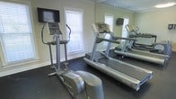 This fitness room is thoughtfully designed to accommodate a focused cardio workout experience within a single-floor layout. The space is equipped with key exercise machines, including a treadmill and an elliptical trainer, both from recognizable commercial-grade brands. A large floor-to-ceiling mirror lines one wall, effectively doubling the perceived size of the room and allowing users to monitor their form during workouts. Wall-mounted flat-screen televisions are positioned for easy viewing from both machines, adding an entertainment element to exercise sessions. Natural light filters in through multiple windows fitted with white blinds, creating a bright and motivating atmosphere. The dark rubberized flooring provides both safety and durability, ideal for a high-use fitness environment. Crown molding along the ceiling adds a subtle architectural detail that elevates the room's finish. The overall layout is efficient, maximizing the available floor space while keeping the environment clean and uncluttered, making it a practical and well-appointed fitness area.